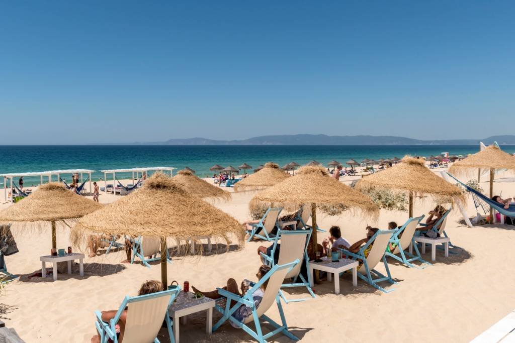 Foto colorida mostra uma praia com guarda-sói sde palha num dia ensolarado