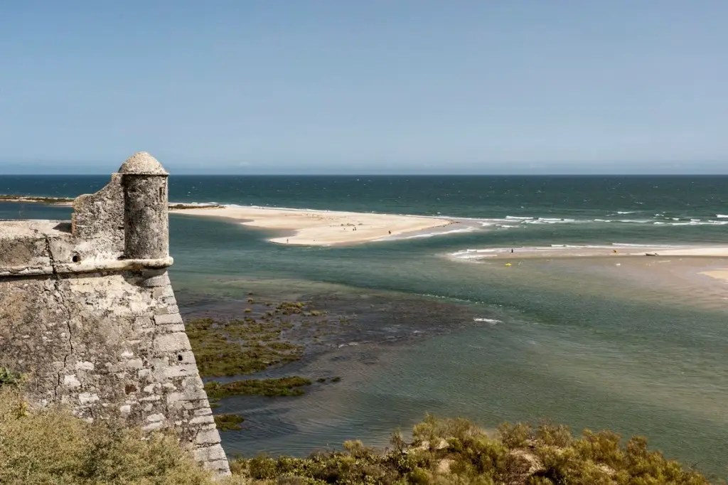 Foto colorida mostra uma praia vista do alto de uma muralha, com águas azuis e verdes e bancos de areia