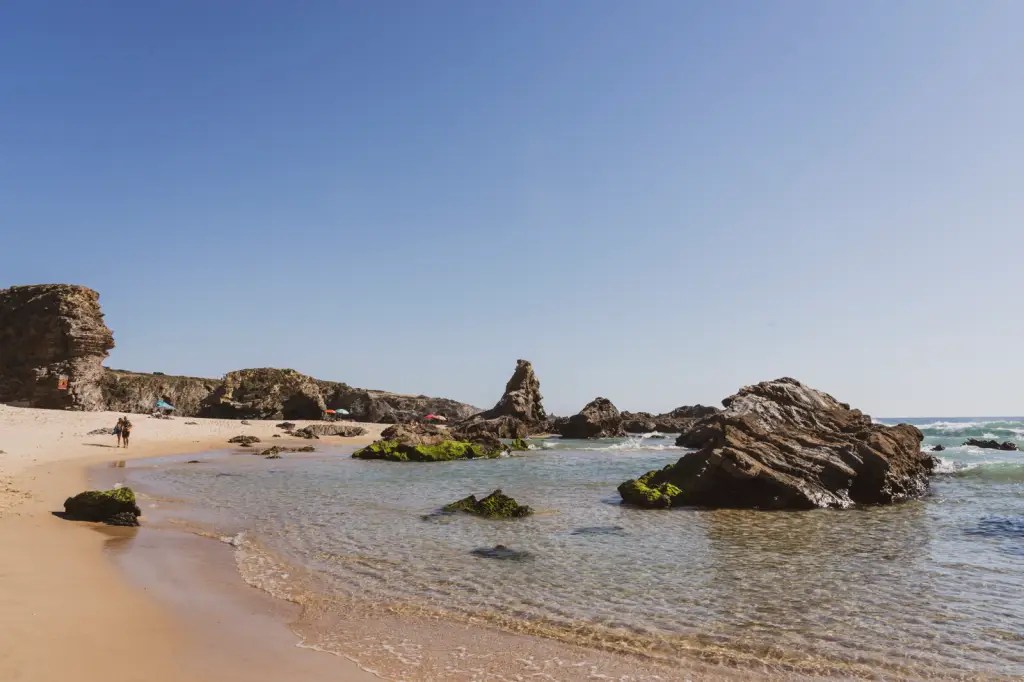 Foto colorida mostra uma praia de águas transparentes e pedras que emergem do mar