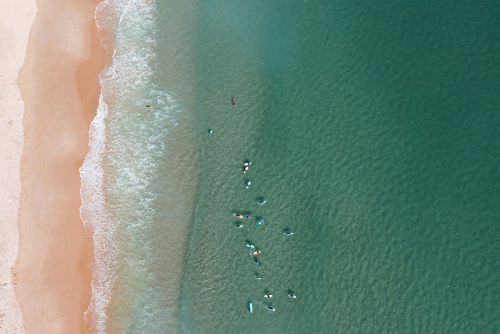 Imagem aérea colorida mostra uma praia com o mar cheio de surfistas a esperar por uma onda