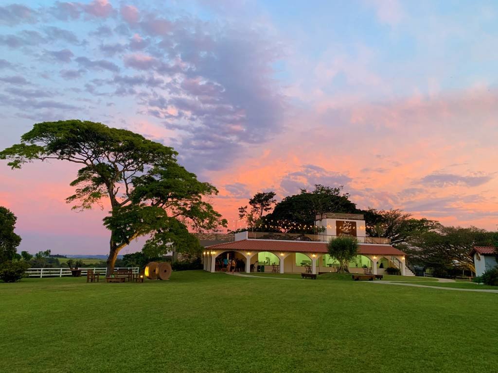 Na imagem vemos uma construção, que parece uma casa grande, em meio à um campo verde.