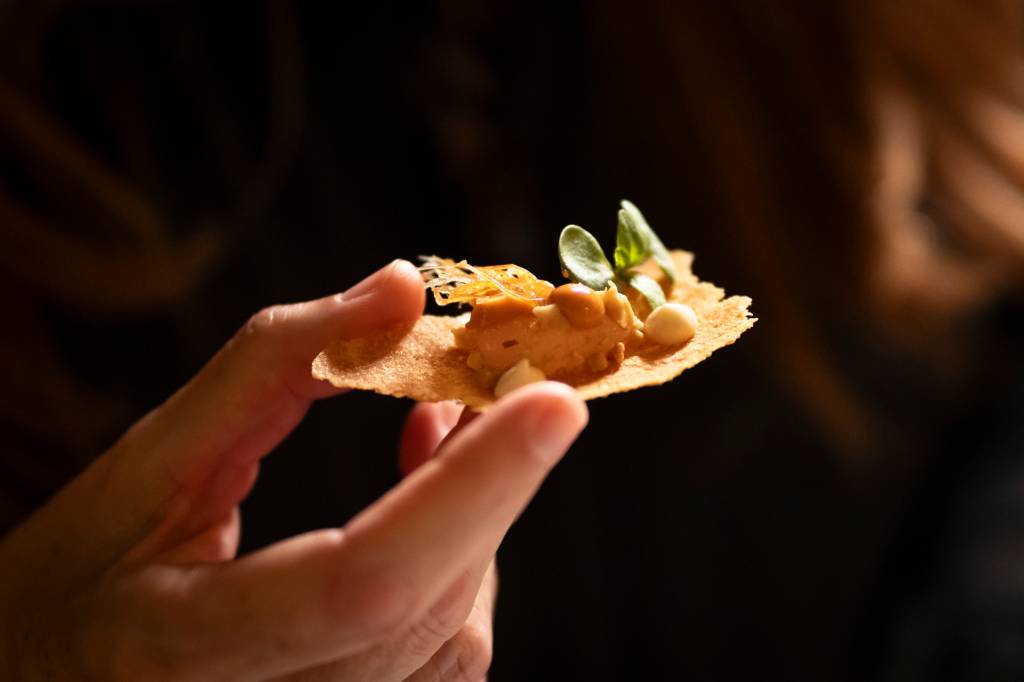 A imagem mostra um dos petiscos do restaurante na mão de uma cliente, à frente de um fundo preto, onde se vê uma casquinha crocante e algumas folhas