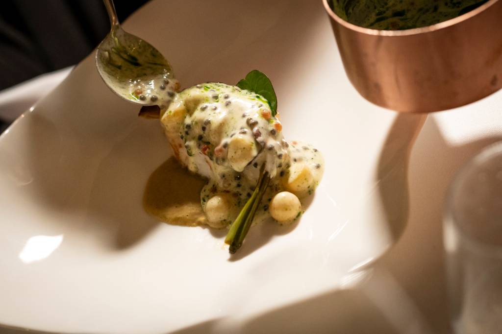 Um prato do restaurante: Ao centro de um prato de louça branca vê-se um peixe e alguns legumes, com uma panelinha de cobre de onde se vê uma colher a retirar o molho