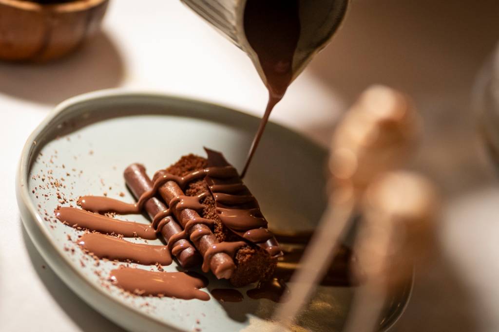 Uma sobremesa do restaurante, onde se vê diferentes texturas de chocolate, com uma calda caindo sobre elas